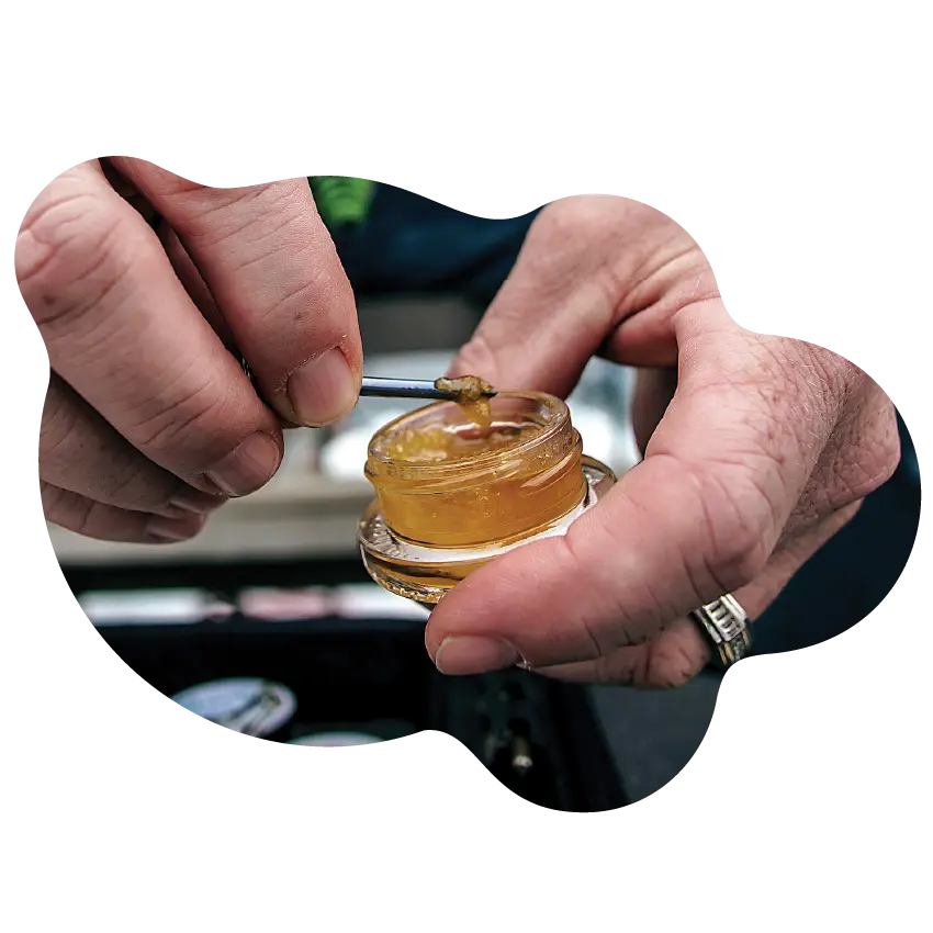 Close-up of hands using a dab tool to scoop golden cannabis concentrate from a small glass jar, featuring a product available at Housing Works Cannabis Co. dispensary.