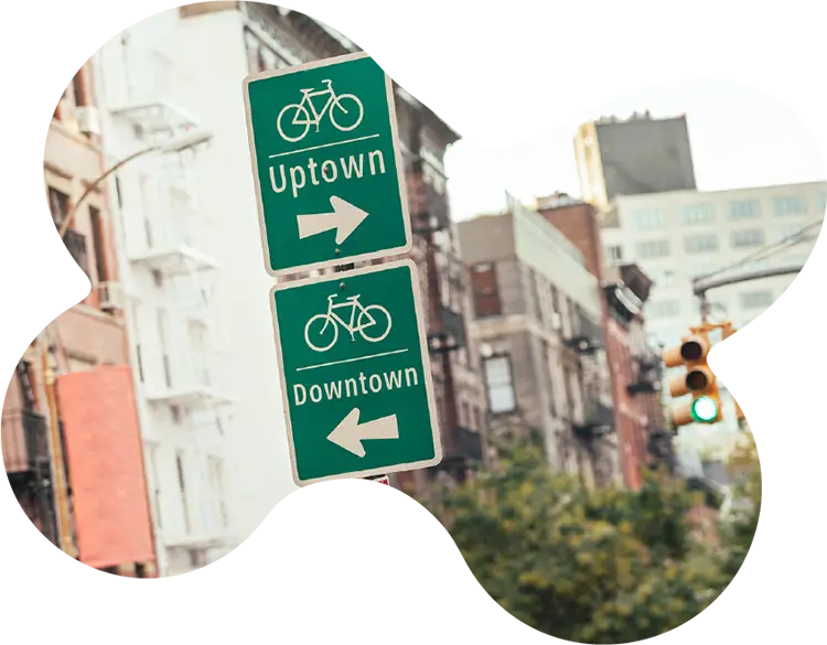 Green bike lane signs in New York City indicating directions for Uptown (right) and Downtown (left), with urban buildings and a traffic light in the background.