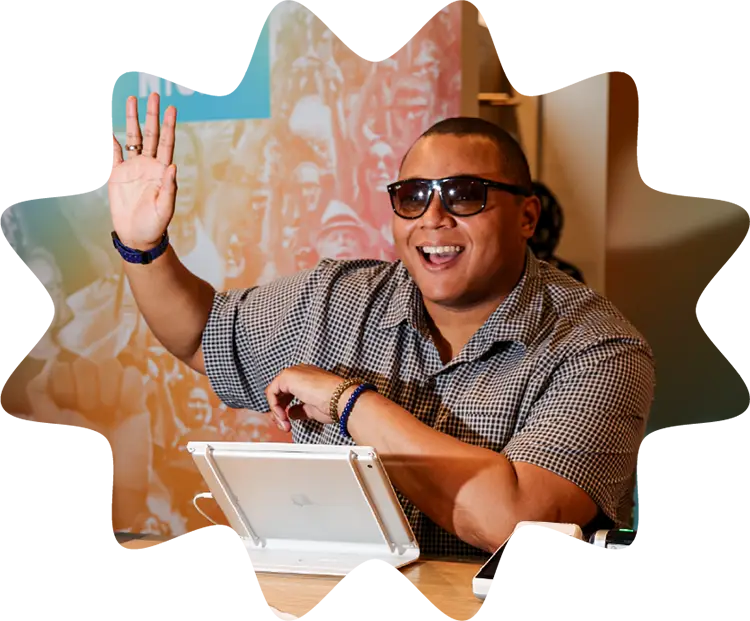 Smiling Housing Works Cannabis Co. staff member wearing sunglasses and raising a hand while seated at a customer service desk.